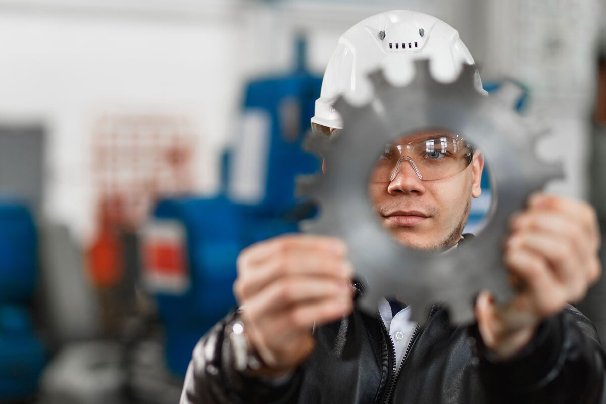 Worker in hat and googles visually inspecting metal cog